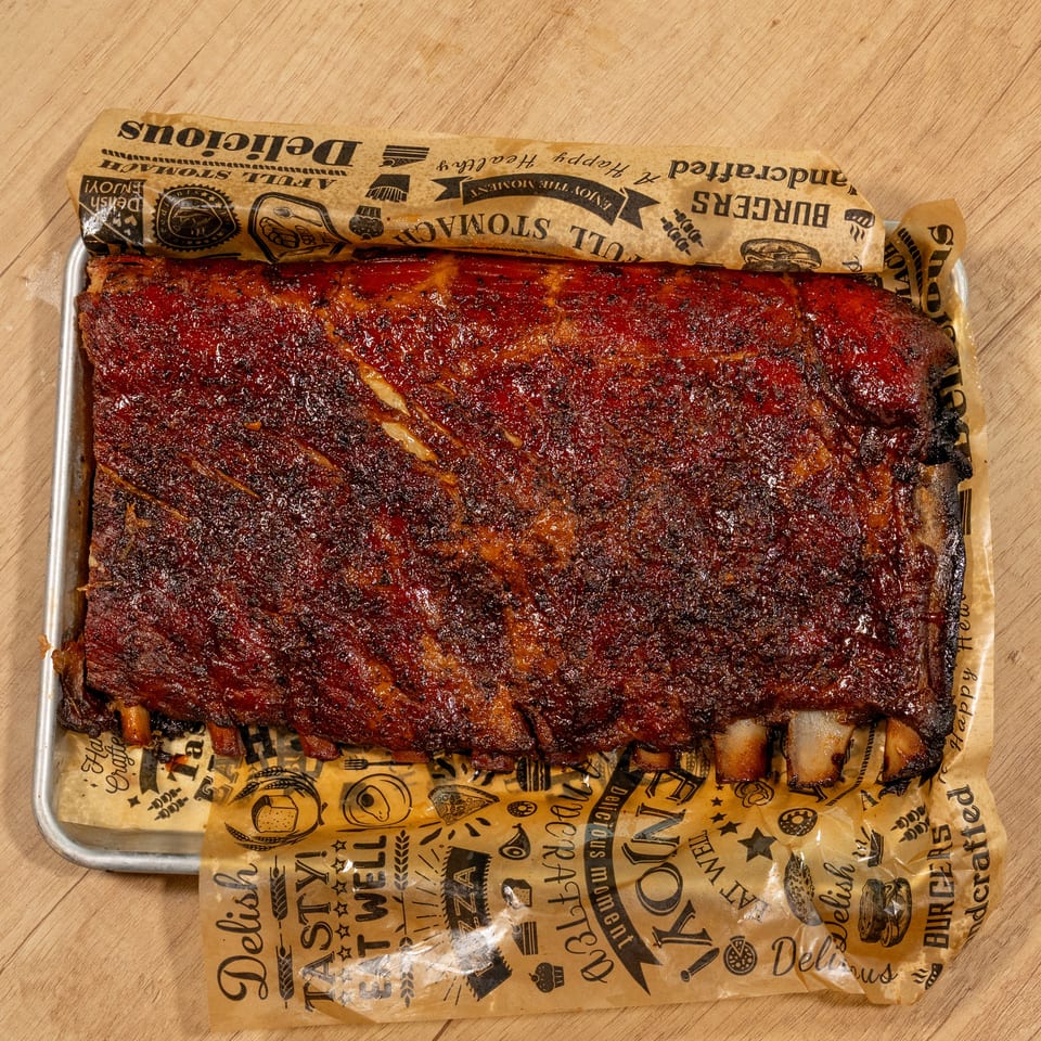 A full rack of glistening, saucy BBQ ribs on a metal tray lined with branded paper.