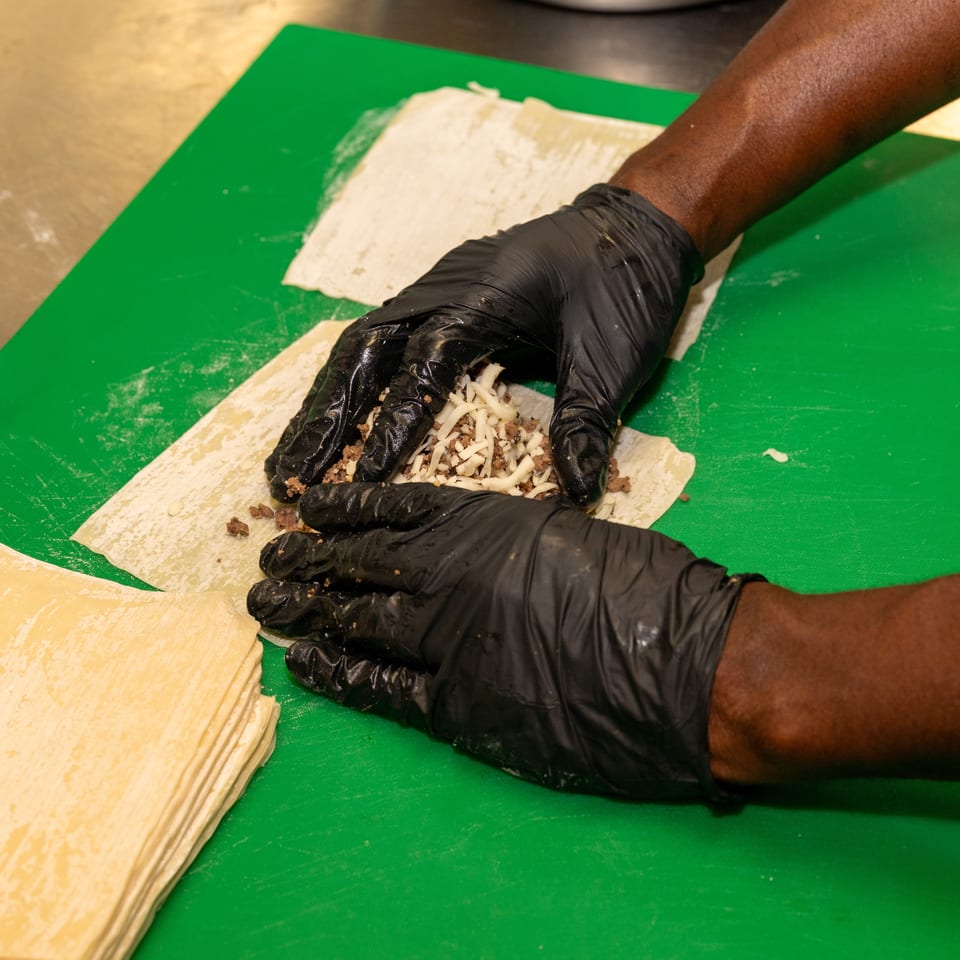Gloved hands preparing egg rolls with ground meat and shredded cheese on a green cutting board.