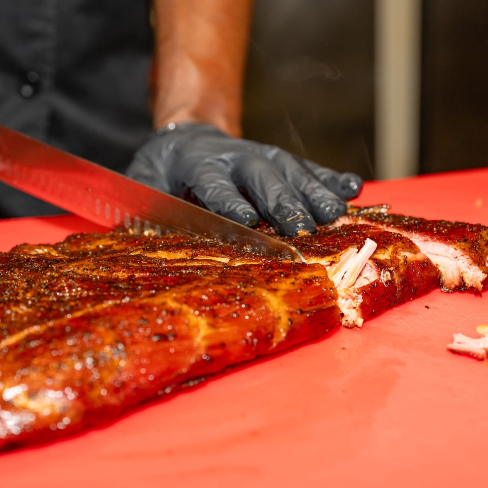 Chef in black gloves slicing a rack of glistening, smoked ribs on a red cutting board.