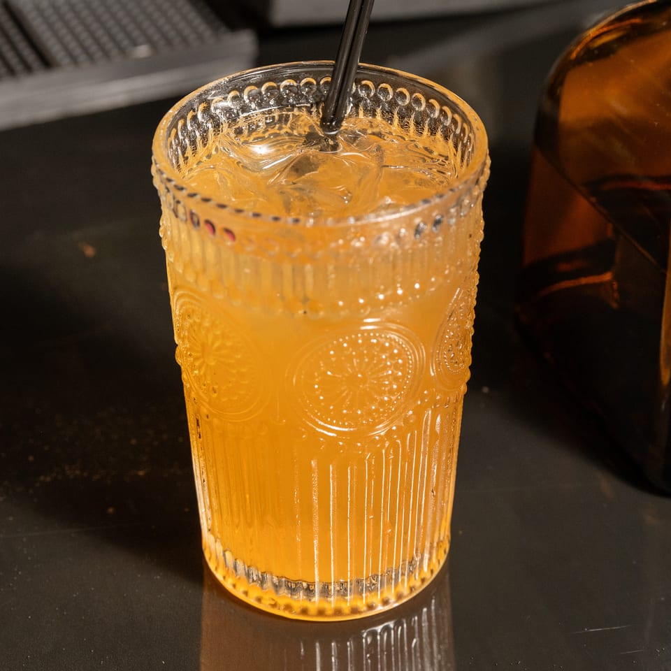 Orange cocktail with ice and a black straw in a textured glass on a dark bar.