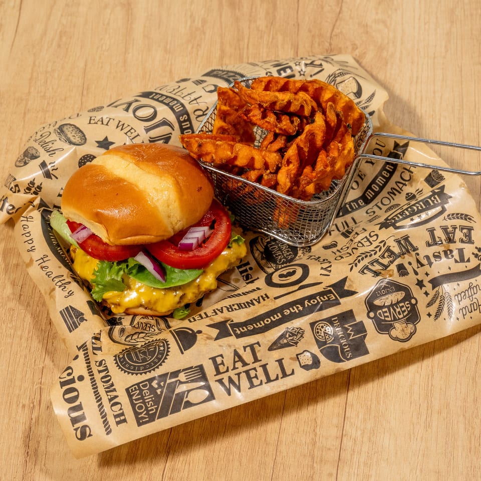 Cheeseburger with lettuce, tomato, and onion, served with waffle-cut sweet potato fries on branded paper.