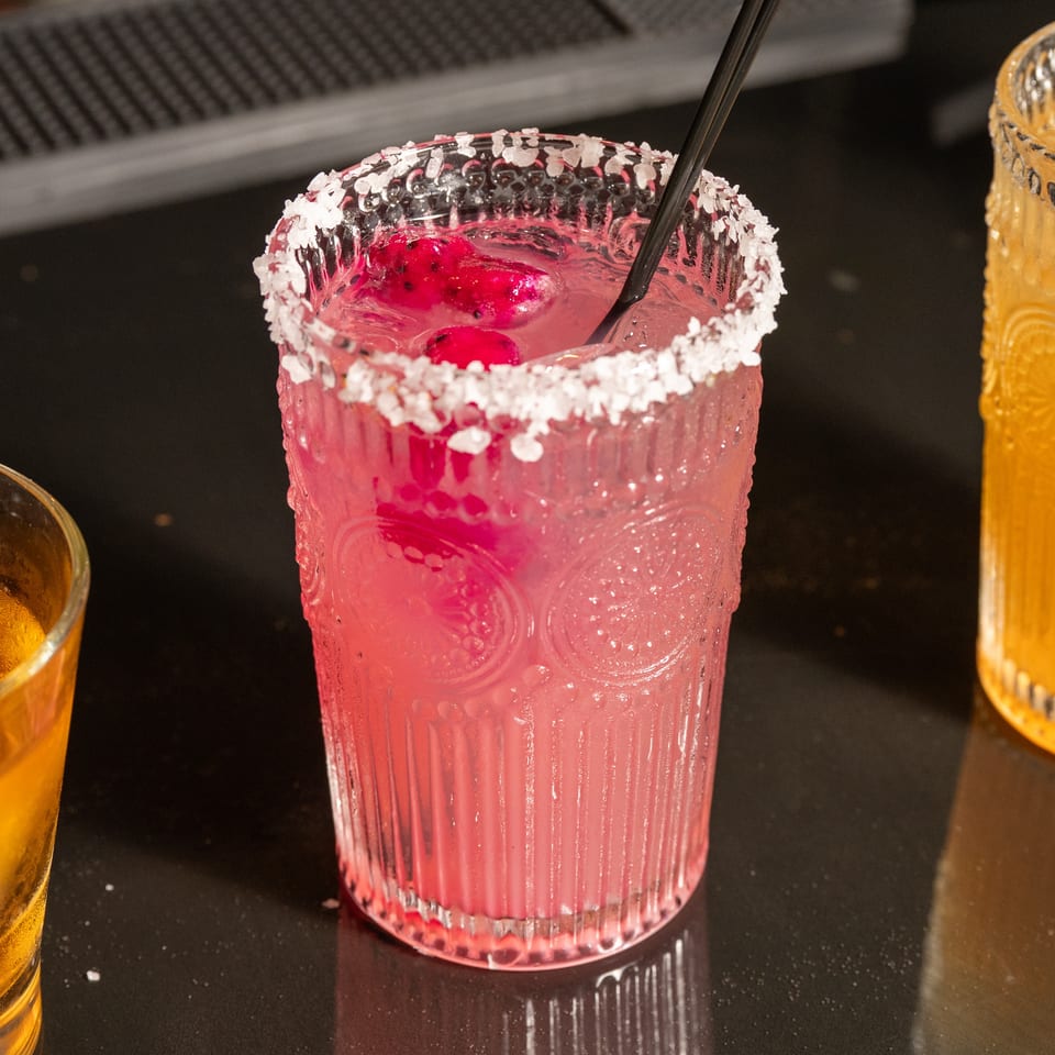 A vibrant pink cocktail with dragon fruit and a salted rim, alongside other drinks on a dark bar.